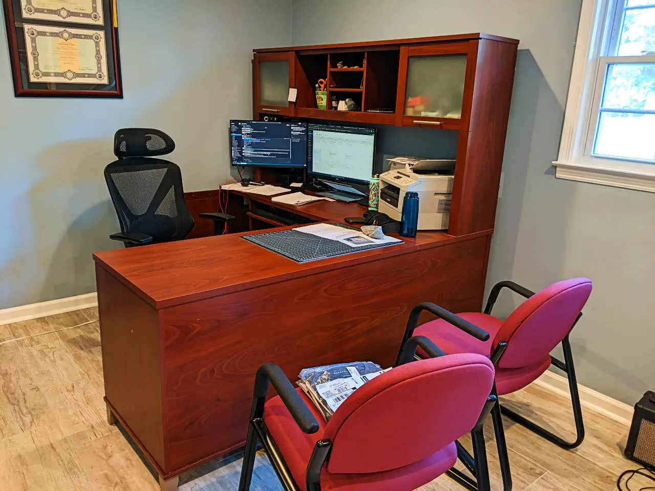 La scrivania con computer, stampante, due schermi e le lauree appese al muro