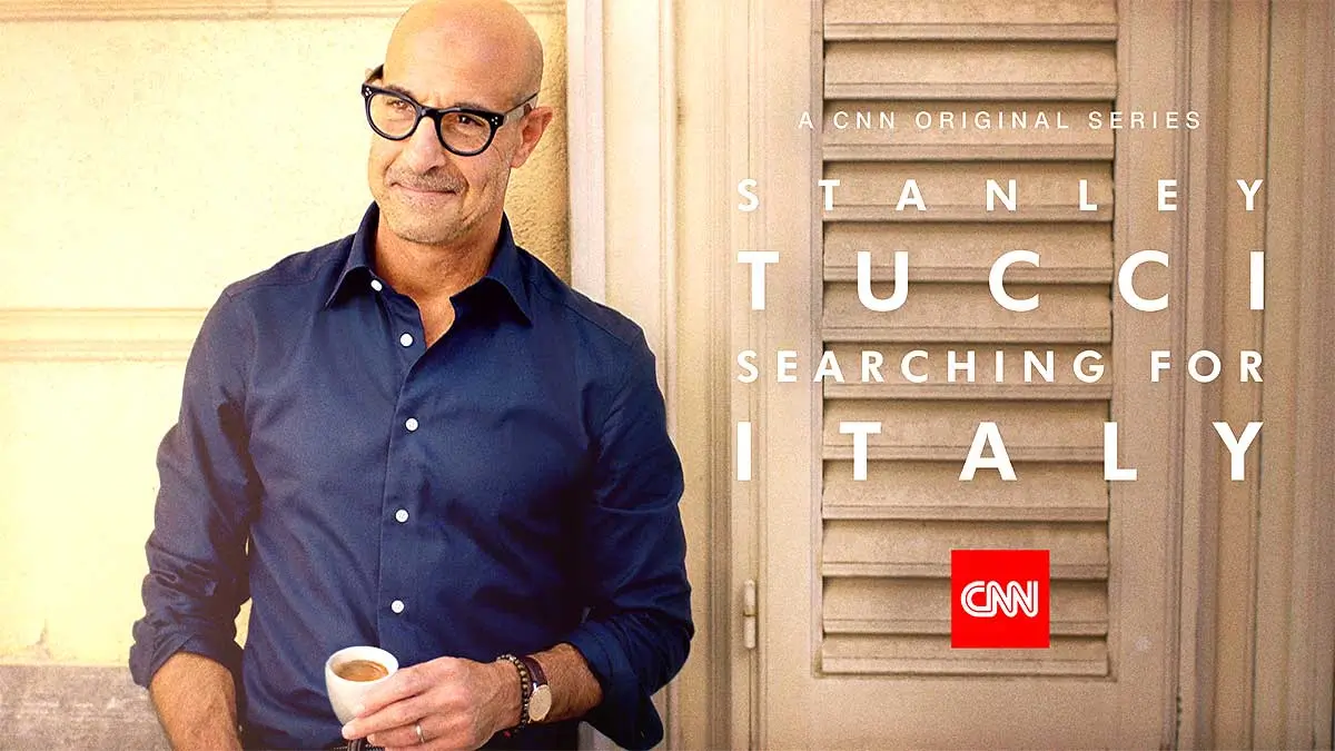 Stanley Tucci sorridente vicino ad una persiana mentre tiene in mano una tazzina di espresso